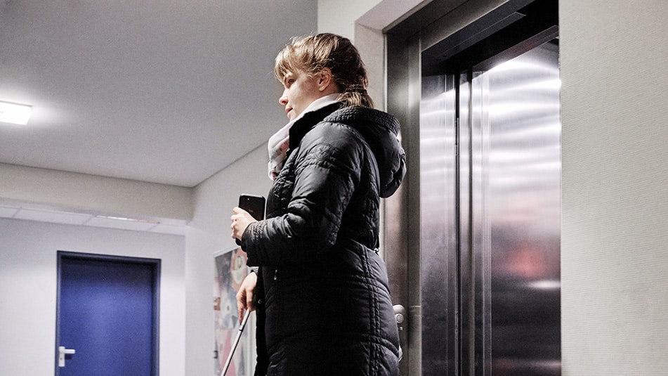 A woman who is blind exiting an elevator using the Blindsquare application on her mobile phone and her white cane.
